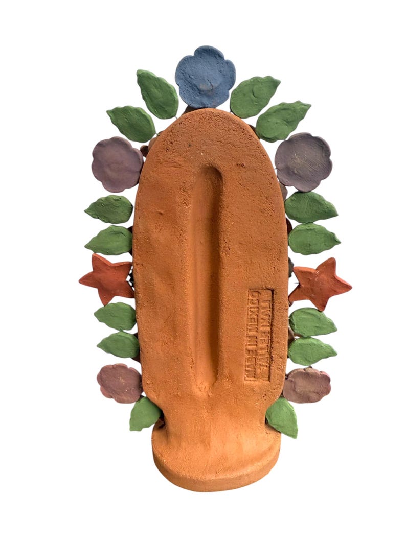 May include: A terracotta-colored, handcrafted decorative piece with a central, elongated shape. It is adorned with colorful, stylized flowers, leaves, and stars in shades of blue, green, purple, and orange. The text "MADE IN MEXICO" is visible.