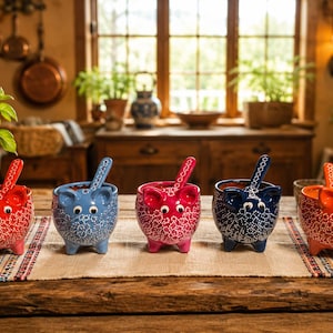 May include: Five ceramic pig-shaped bowls with matching spoons, each featuring a distinct solid color—red, light blue, pink, dark blue, and orange—adorned with a white floral pattern, small ears, and two eyes on the front of each bowl.