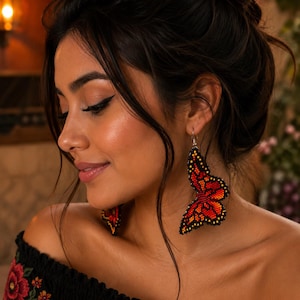 Op de afbeelding: Een vrouw draagt ​​geparelde vlinder oorbellen. De oorbellen zijn voornamelijk rood en oranje met zwarte accenten en hangen aan zilveren haken. De vrouw draagt ​​een zwarte off-the-shoulder top met bloemenborduurwerk.