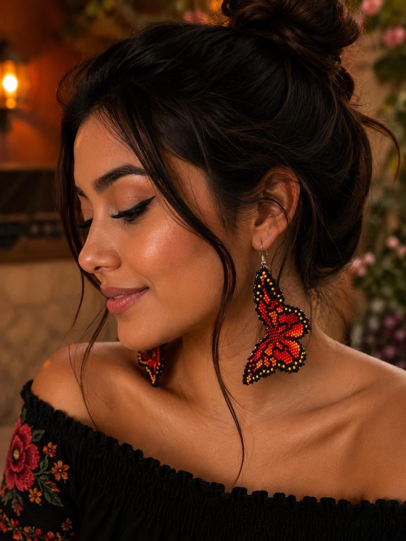 Op de afbeelding: Een vrouw draagt ​​geparelde vlinder oorbellen. De oorbellen zijn voornamelijk rood en oranje met zwarte accenten en hangen aan zilveren haken. De vrouw draagt ​​een zwarte off-the-shoulder top met bloemenborduurwerk.