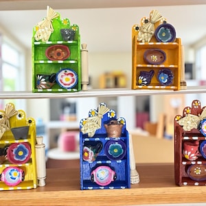 Può includere: Mini scaffali in legno dipinti a mano in verde, giallo, blu e rosso, ciascuno con piccoli piatti e tazze decorative. Gli scaffali hanno un design rustico con accenti di paglia e dettagli floreali, perfetti per una casa delle bambole.
