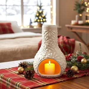 Peut inclure: Un bougeoir en céramique blanc orné de motifs floraux, contenant une bougie jaune allumée. Il est posé sur un chemin de table à carreaux rouges, décoré de pommes de pin et de boules. L'ensemble évoque une ambiance chaleureuse et festive.