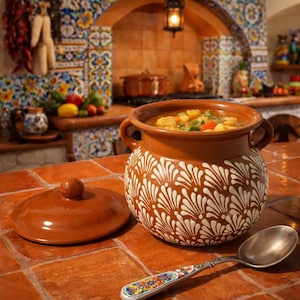 May include: A brown ceramic soup pot with a white floral pattern, filled with soup. The pot is on a terracotta tile surface next to its lid and a decorative spoon. The background shows a kitchen with colourful tilework.