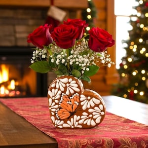 Puede incluir: Un jarrón en forma de corazón con un diseño de mariposa contiene rosas rojas y flores blancas. El jarrón marrón tiene detalles blancos y naranjas. El arreglo está sobre un camino de mesa rojo.