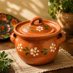 May include: A round terracotta cooking pot with two side handles and a matching lid. The exterior of the pot and the lid are decorated with hand-painted white flowers featuring yellow centres and green leaves against the natural reddish-brown clay.
