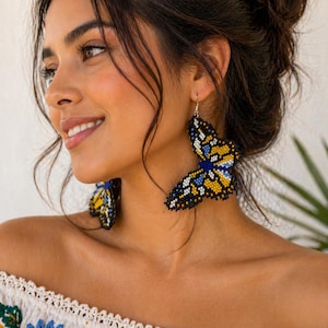 Op de afbeelding: Vlinder-vormige oorbellen van blauwe, gele en zwarte kralen. De oorbellen hangen aan zilveren haken. Een uniek accessoire, perfect om een vleugje fantasie aan elke outfit toe te voegen.