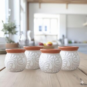 May include: Four small, white ceramic pots with a floral design and a terracotta-colored rim. The pots are arranged on a light-colored wooden table. The background is a blurred kitchen scene.