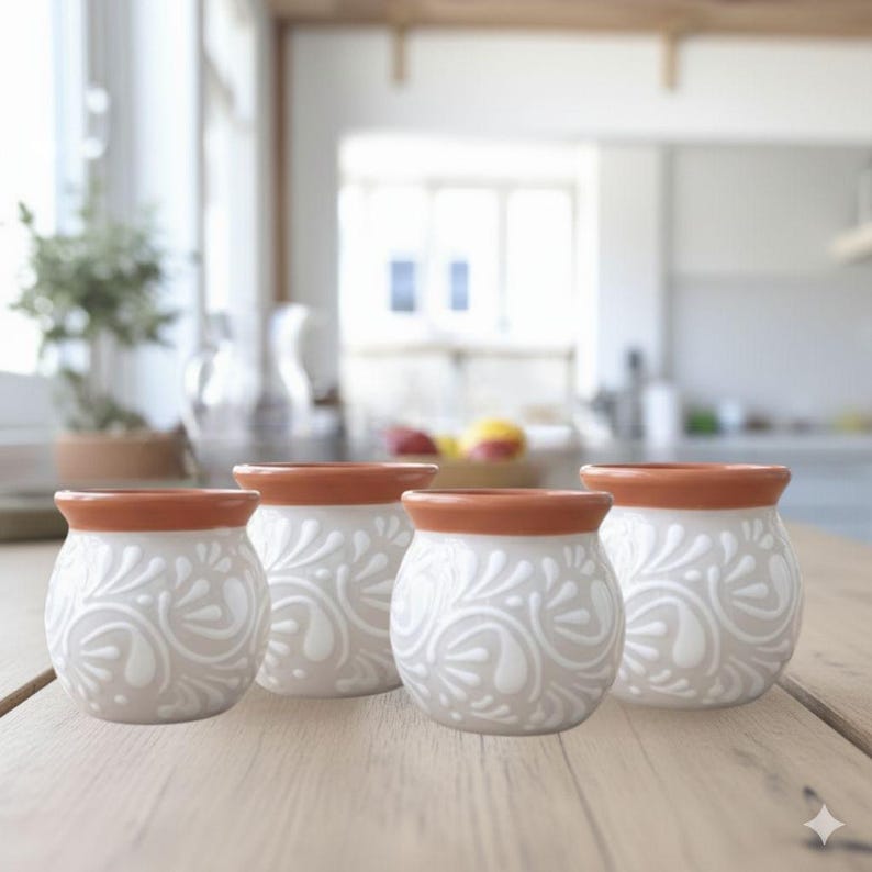 May include: Four small, white ceramic pots with a floral design and a terracotta-colored rim. The pots are arranged on a light-colored wooden table. The background is a blurred kitchen scene.