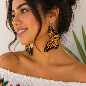 Op de afbeelding: Vlinder-vormige oorbellen van oranje, gele en zwarte kralen. De oorbellen hangen aan zilveren haken. De vrouw draagt een witte off-the-shoulder top met kleurrijke bloemenborduursels.