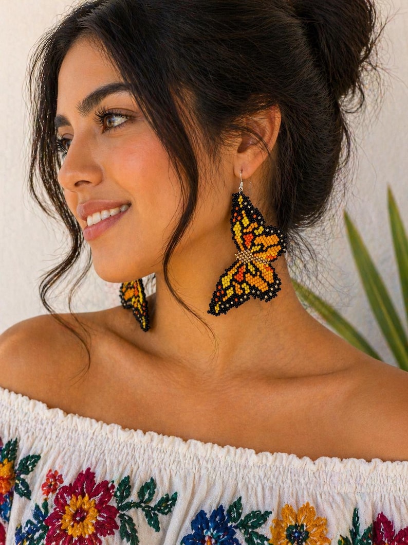 Op de afbeelding: Vlinder-vormige oorbellen van oranje, gele en zwarte kralen. De oorbellen hangen aan zilveren haken. De vrouw draagt een witte off-the-shoulder top met kleurrijke bloemenborduursels.