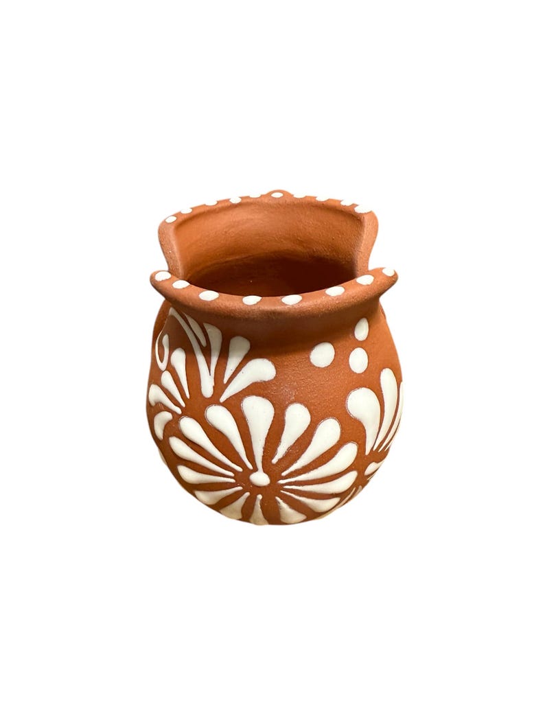 Op de afbeelding: Een kleine terracotta-kleurige keramische pot met een wit bloemmotief. De pot heeft een afgeronde body en een uitlopende rand met witte stippen. Het witte bloemmotief is symmetrisch en bedekt het grootste deel van het oppervlak van de pot.
