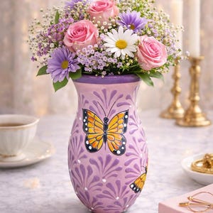 May include: A lavender ceramic vase with a butterfly design holds a bouquet of pink roses, daisies, and purple flowers. The vase is decorated with a repeating leaf pattern and a yellow and black butterfly. Two lit candles are in the background.