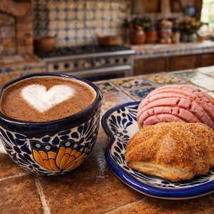May include: A cup of coffee with a heart-shaped design in the foam, next to a plate with two pastries. The cup has a blue and white floral pattern with an orange butterfly. The pastries are a pink concha and a golden-brown pastry.