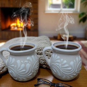 Może przedstawiać: Dwa białe ceramiczne kubki wypełnione kawą, ozdobione kwiatowym wzorem, stoją na drewnianej powierzchni. Para okularów w czarnych oprawkach leży obok. Scena przywołuje ciepło i komfort.