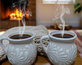 May include: Two white ceramic mugs filled with dark coffee, adorned with a floral design, sit on a wooden surface. Steam rises from the coffee. A pair of black-framed glasses rests nearby. The scene evokes warmth and comfort.