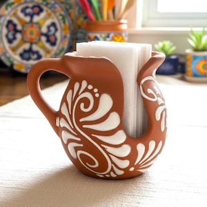 Op de afbeelding: Een terracotta servethouder met een handvat en een gebogen ontwerp. Witte bloempatronen sieren het oppervlak van de houder. Het bevat een stapel witte servetten, ideaal voor keuken- of eetkamerdecoratie.