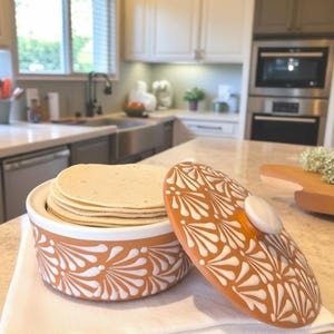 Pode incluir: Um aquecedor de tortillas de cerâmica com tampa, cheio de tortillas. O aquecedor laranja e branco está sobre um pano branco. A tampa tem um design correspondente. O fundo da cozinha inclui uma janela, uma pia e armários.