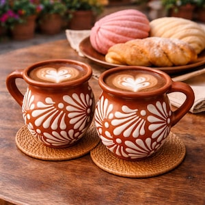 Pode incluir: Duas canecas de terracota com desenhos florais brancos, cheias de café e latte art em forma de coração, estão sobre bases tecidas. Um prato de doces, incluindo conchas, no fundo, sugerindo um ambiente de café.