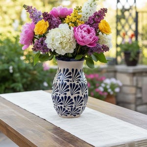 May include: A colourful flower arrangement in a patterned vase. The bouquet includes pink peonies, yellow roses, white hydrangeas, and purple lilacs. The vase is cream with a blue design, placed on a wooden table with a cream table runner.