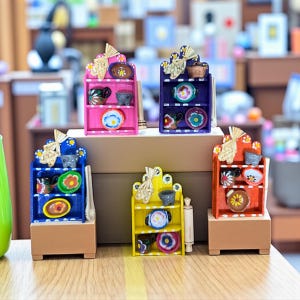 Può includere: Piccoli scaffali in legno rosa, blu, giallo e arancione, ciascuno con minuscoli piatti e tazze decorativi. Gli scaffali hanno una parte superiore decorativa e sono posizionati su una base di legno. Un'esposizione affascinante e colorata.