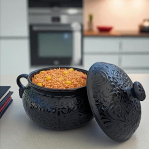 May include: A black ceramic serving bowl with a lid, filled with rice and corn. The bowl and lid feature an embossed floral design. The lid is partially open, revealing the contents. The bowl has two handles.