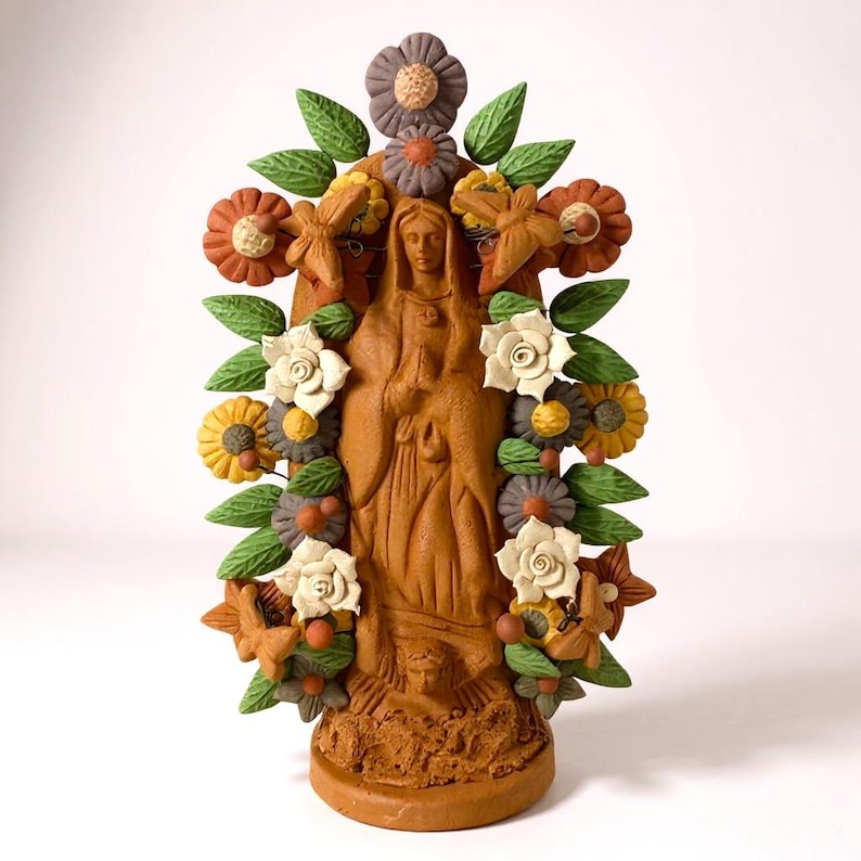 May include: A terracotta-colored statue of a religious figure, adorned with a floral wreath of green leaves, white, yellow, and orange flowers. The statue is standing with hands clasped in prayer.