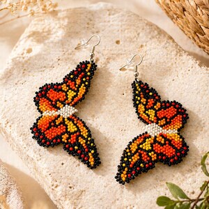 Op de afbeelding: Een paar oorbellen in de vorm van een vlinder, gemaakt van kralen. De oorbellen hebben oranje, gele en zwarte kralen, met een wit kralen midden. Ze hebben zilverkleurige haken.