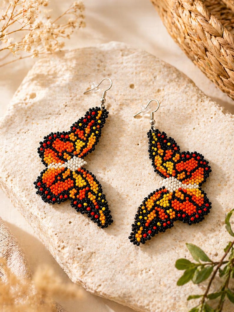 Op de afbeelding: Een paar oorbellen in de vorm van een vlinder, gemaakt van kralen. De oorbellen hebben oranje, gele en zwarte kralen, met een wit kralen midden. Ze hebben zilverkleurige haken.