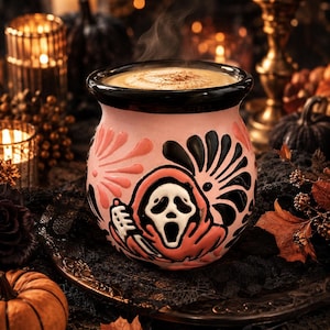 Puede incluir: Una taza redonda de cerámica rosa con borde negro, que presenta un diseño en relieve de una figura con máscara blanca y túnica con capucha que sostiene un cuchillo, rodeada de patrones florales negros y rosas, llena de una bebida humeante.
