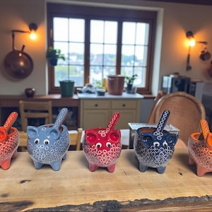 May include: Five ceramic pig-shaped bowls with spoons, each in a different color: red, blue, red, dark blue, and orange. The bowls have white floral patterns and are displayed on a wooden surface. The spoons match the bowl colors.