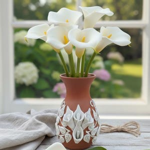 Pu&ograve; includere: Un vaso in terracotta con calle bianche. Il vaso presenta un motivo floreale bianco decorativo. Una singola calla &egrave; appoggiata sulla superficie di legno. Lo sfondo include una finestra con vista sul verde e un panno colorato neutro.