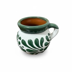 May include: A handcrafted ceramic mug with a white base, adorned with green floral patterns. The rim and handle are green, and the interior is a warm brown. This traditional mug is perfect for serving hot beverages.