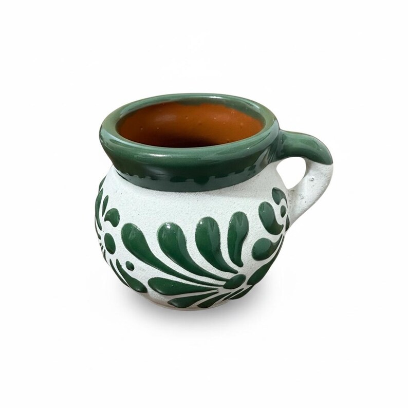May include: A handcrafted ceramic mug with a white base, adorned with green floral patterns. The rim and handle are green, and the interior is a warm brown. This traditional mug is perfect for serving hot beverages.
