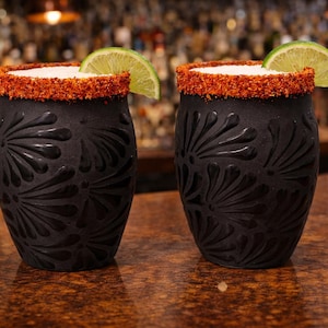 Può includere: Due bicchieri da cocktail in ceramica nera con un motivo floreale strutturato, riempiti con una bevanda bianca e guarniti con una fetta di lime. I bordi sono ricoperti da una spezia rossa. I bicchieri sono su una superficie marrone.