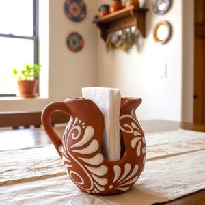 Puede incluir: Un servilletero de terracota con diseños florales blancos. El soporte contiene servilletas blancas y está colocado sobre una mesa de madera. El fondo incluye una ventana, platos decorativos y una estantería con plantas.