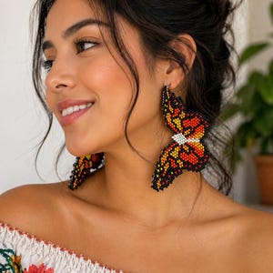 Op de afbeelding: Vlinder-vormige kralen oorbellen in zwart, oranje, geel en wit. De oorbellen zijn een gedetailleerde weergave van een vlinder, met ingewikkeld kralenwerk. Een uniek accessoire.