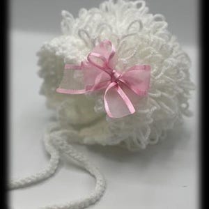 Könnte beinhalten: Eine weiße, flauschige Babyhaube mit einer rosa Schleife. Die Haube besteht aus einem weichen, strukturierten Material und hat zwei weiße Bänder. Die Haube ist für Säuglinge und Kleinkinder konzipiert.