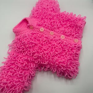 May include: A vibrant pink, hand-crocheted sweater with a distinctive looped texture. The sweater has a button-up front with four clear, flower-shaped buttons. The sleeves are long, and the design is cosy and textured.