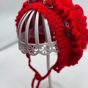 May include: Red crocheted baby bonnet with a ruffled edge and three red fabric roses with green leaves. The bonnet is on a white display stand.