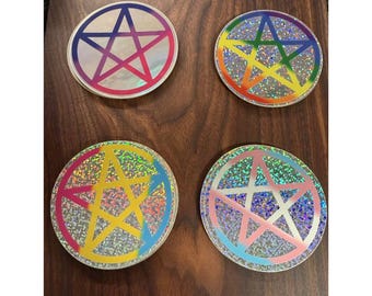 May include: Four round coasters with a pentagram design. Each coaster features a different color scheme: red and pink, rainbow, yellow and pink, and pink and blue. The coasters have a glittery, iridescent background.