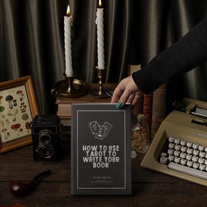Pode incluir: Um livro intitulado "How to Use Tarot to Write Your Book" com ilustra&ccedil;&otilde;es de cartas de tar&ocirc;. O livro est&aacute; em uma mesa de madeira com uma m&aacute;quina de escrever vintage, uma impress&atilde;o emoldurada de cogumelos, velas e uma c&acirc;mera vintage.