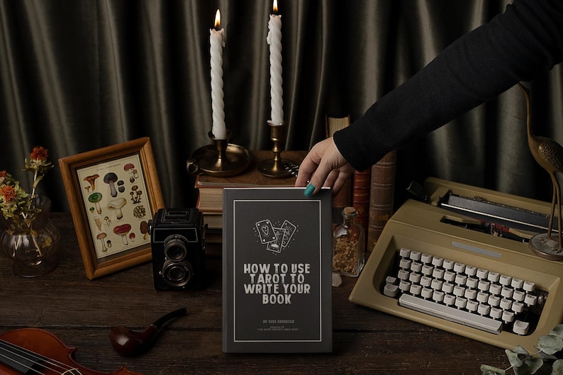 Pode incluir: Um livro intitulado "How to Use Tarot to Write Your Book" com ilustra&ccedil;&otilde;es de cartas de tar&ocirc;. O livro est&aacute; em uma mesa de madeira com uma m&aacute;quina de escrever vintage, uma impress&atilde;o emoldurada de cogumelos, velas e uma c&acirc;mera vintage.