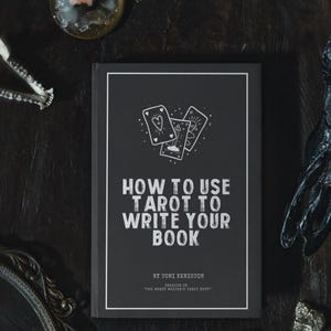 Pode incluir: Um livro intitulado "HOW TO USE TAROT TO WRITE YOUR BOOK" com capa preta e texto branco. A capa apresenta ilustra&ccedil;&otilde;es de cartas de tar&ocirc;. Uma vela acesa e outros objetos m&iacute;sticos circundam o livro em uma superf&iacute;cie escura.