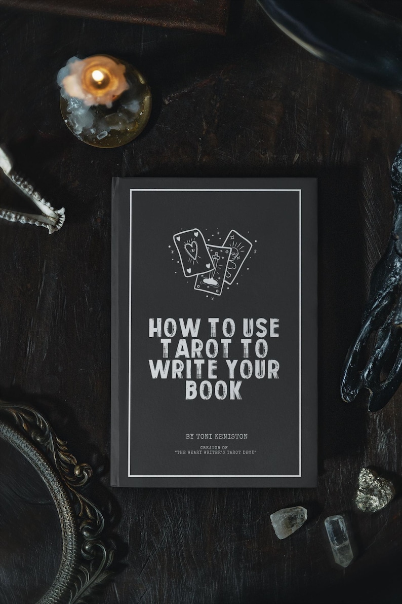 Pode incluir: Um livro intitulado "HOW TO USE TAROT TO WRITE YOUR BOOK" com capa preta e texto branco. A capa apresenta ilustra&ccedil;&otilde;es de cartas de tar&ocirc;. Uma vela acesa e outros objetos m&iacute;sticos circundam o livro em uma superf&iacute;cie escura.