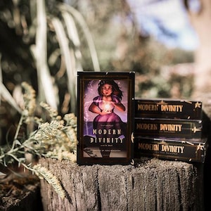 May include: A stack of Modern Divinity tarot card decks. The front deck displays a woman holding a glowing orb. The text "Modern Divinity" is visible on the front of the deck. The decks are black with gold lettering and are stacked on a wooden surface.