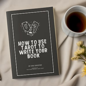 Pode incluir: Um livro intitulado "How to Use Tarot to Write Your Book" com uma capa cinza escuro e texto branco. A capa apresenta ilustra&ccedil;&otilde;es de cartas de tar&ocirc;. Uma caneta branca, uma x&iacute;cara de ch&aacute; e flores s&atilde;o dispostas sobre uma superf&iacute;cie bege.