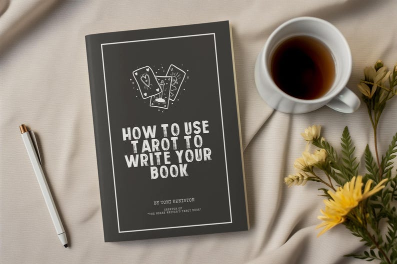 Pode incluir: Um livro intitulado "How to Use Tarot to Write Your Book" com uma capa cinza escuro e texto branco. A capa apresenta ilustra&ccedil;&otilde;es de cartas de tar&ocirc;. Uma caneta branca, uma x&iacute;cara de ch&aacute; e flores s&atilde;o dispostas sobre uma superf&iacute;cie bege.