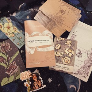 May include: An oracle deck and guidebook titled "Swamp Witch's Oracle" with accompanying cards, a drawstring bag, and stickers. The cards feature botanical illustrations and the guidebook is peach-colored. The items are arranged on a dark background with celestial patterns.