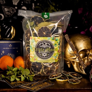 May include: A transparent bag of dried citrus slices, labelled "Triple Moon." The image includes oranges, a golden skull, and a dark blue book. The background features a dark floral pattern.