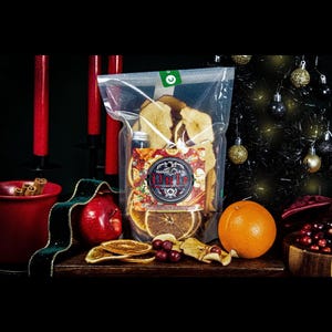 Puede incluir: Una bolsa transparente llena de rodajas de fruta deshidratada, manzanas y naranjas, con una etiqueta que dice "Amaris Oven". Una naranja y una manzana roja están al lado de la bolsa. Velas rojas y una cinta verde están a la izquierda.